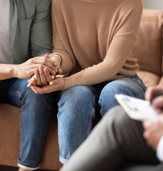 Help Concept. Closeup Cropped View Of Spouses Having Session With Male Marriage Therapist, Sitting On Couch In Office And Holding Each Other Hands. Consultant Writing Taking Notes, Selective Focus